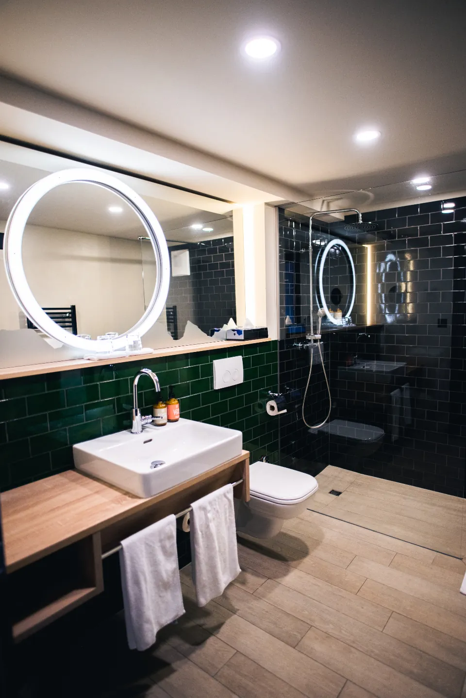 Bathroom with green wall tile and a wood look tile floor under a round lit mirror